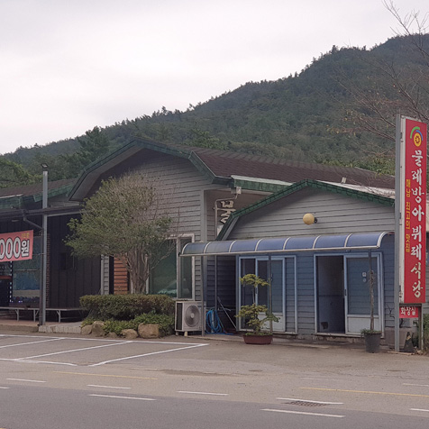 세모모양 초록색 지붕아래 회색 벽면 가운데에 큰 전구가 붙어있고 오른쪽 길가에 '물레방아뷔페식당'이라고 적힌 붉은 간판이 세워져있다